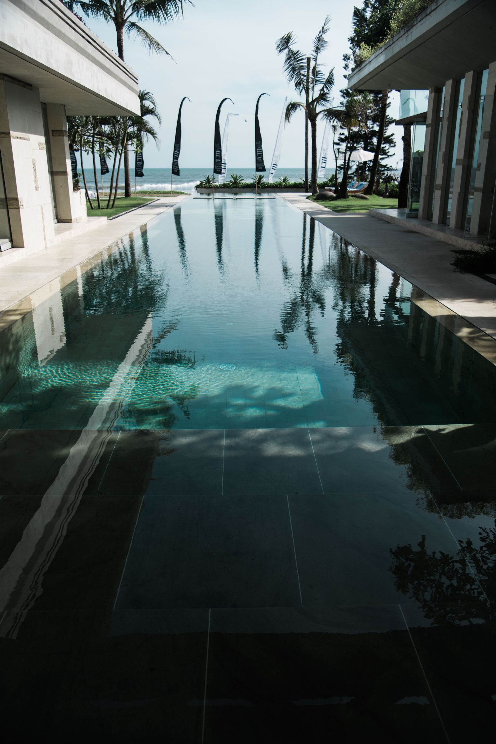 A resort pool framed by palms and buildings opens towards the ocean in Bali.