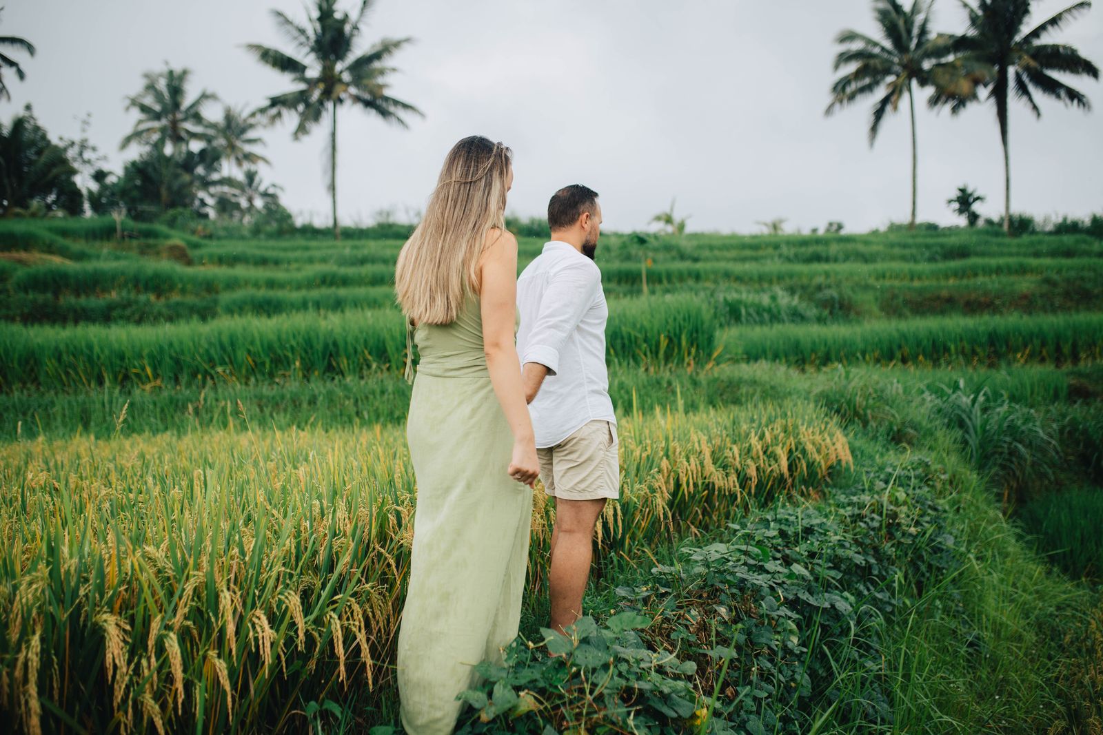A couple holding hands on a narrow path between green rice fields in Bali.