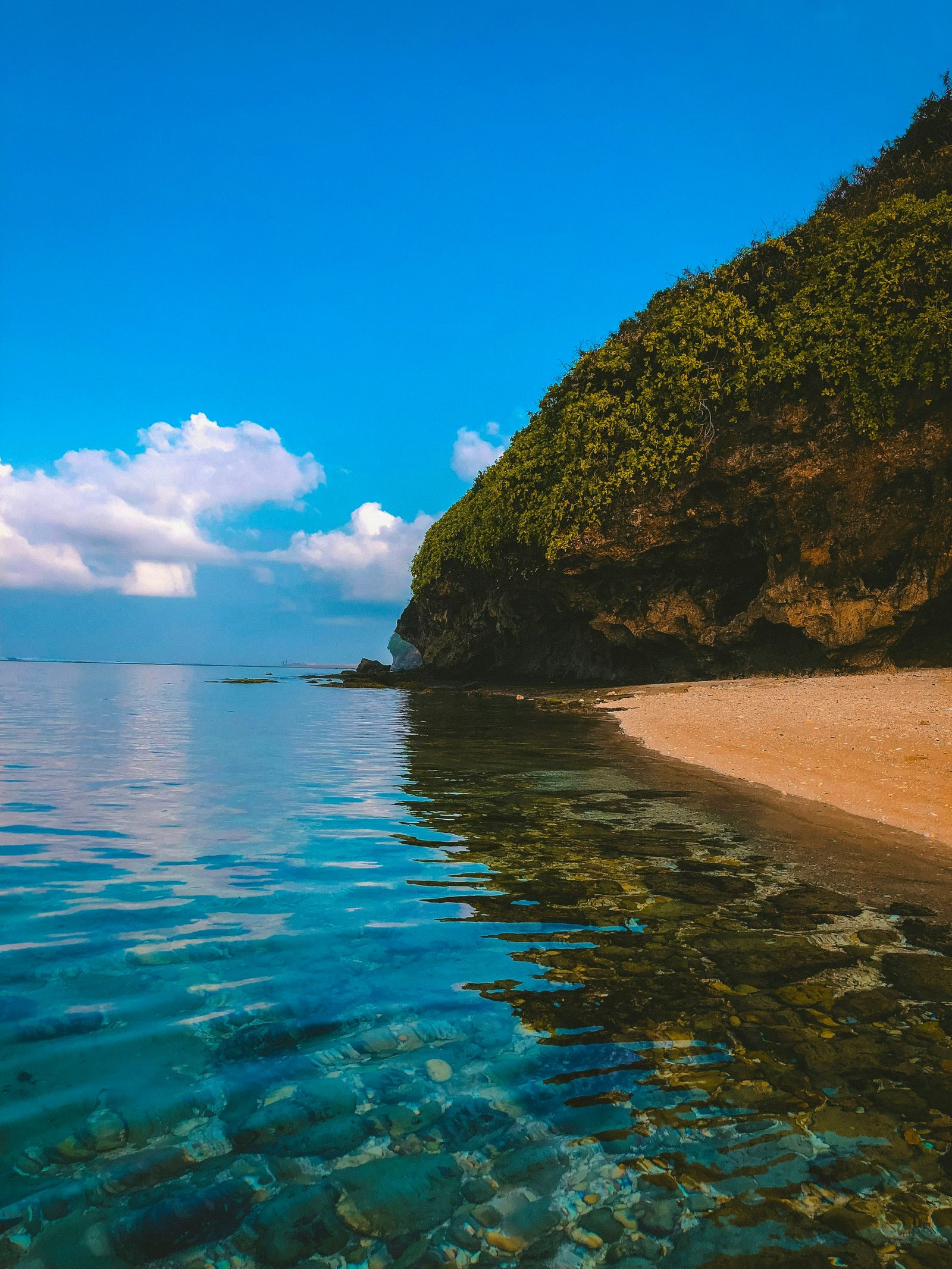 Clear blue water meets a quiet sandy beach beneath a green cliff in Bali.