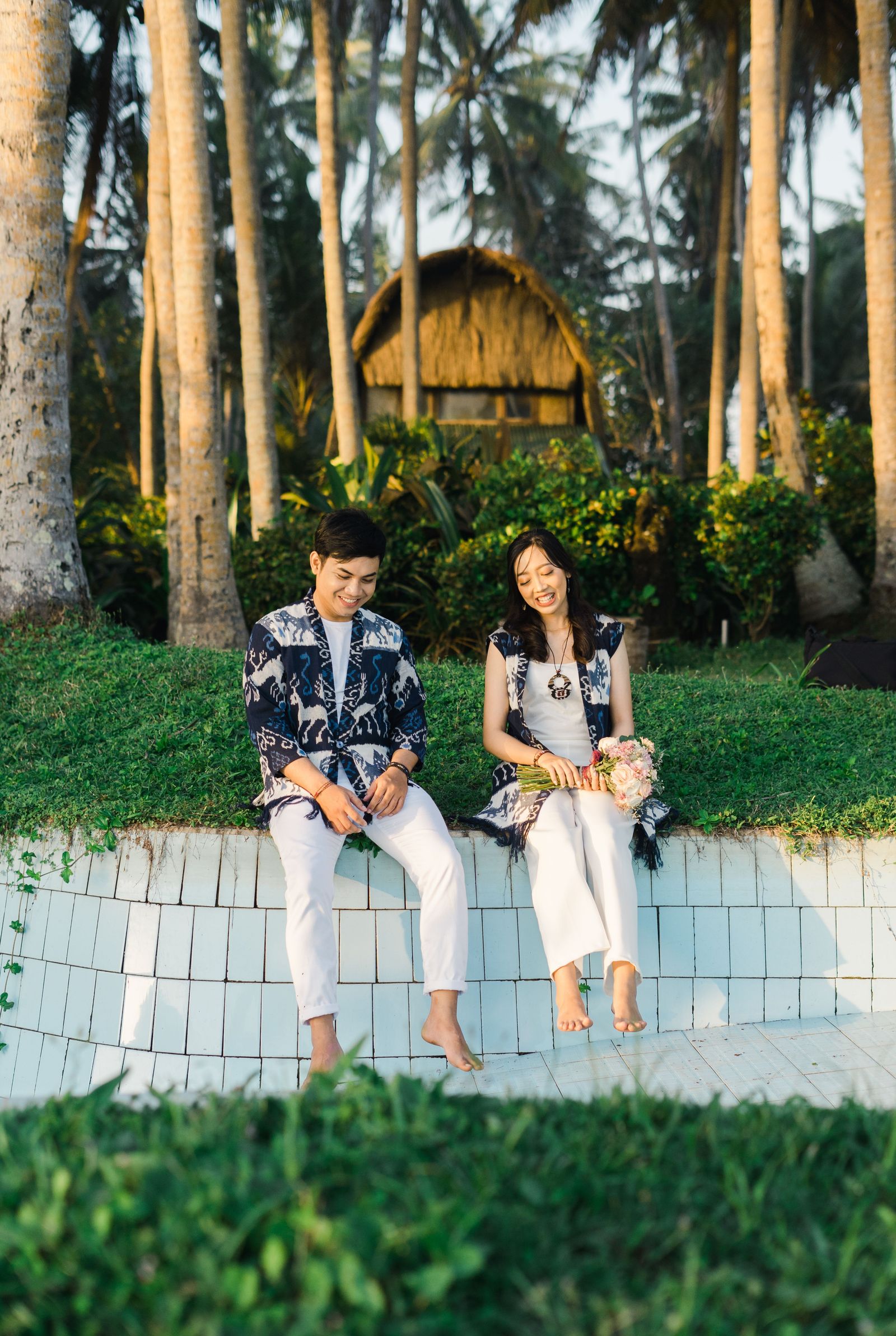 A couple sitting close together and smiling in a tropical garden beneath palm trees.
