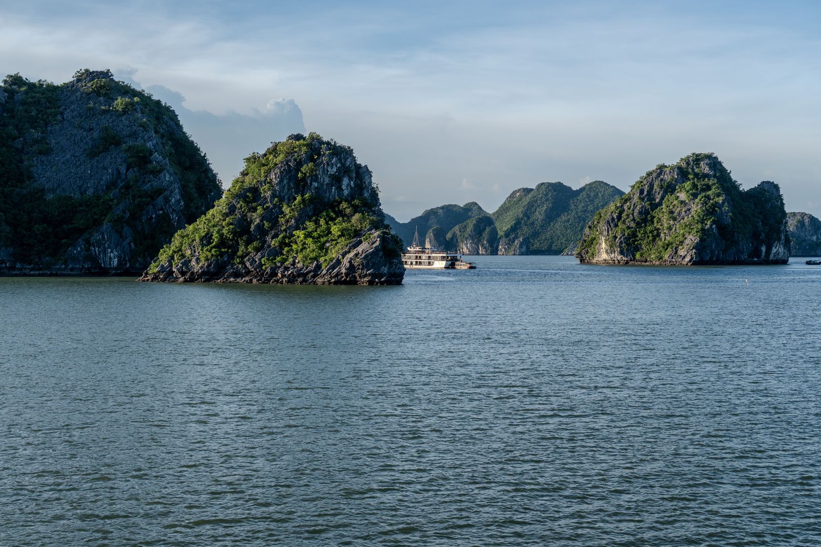 Limestone islands rise from the open water of Ha Long Bay, with small boats in the distance.