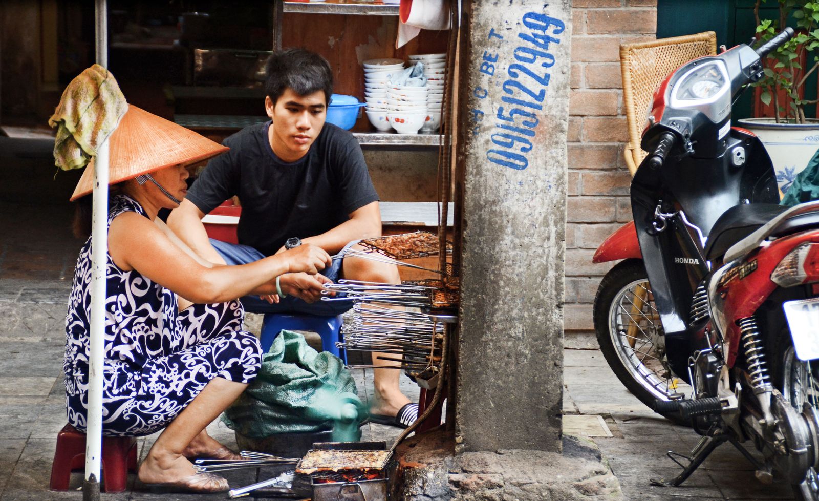 Two street vendors grill food on small charcoal braziers beside a Hanoi pavement.