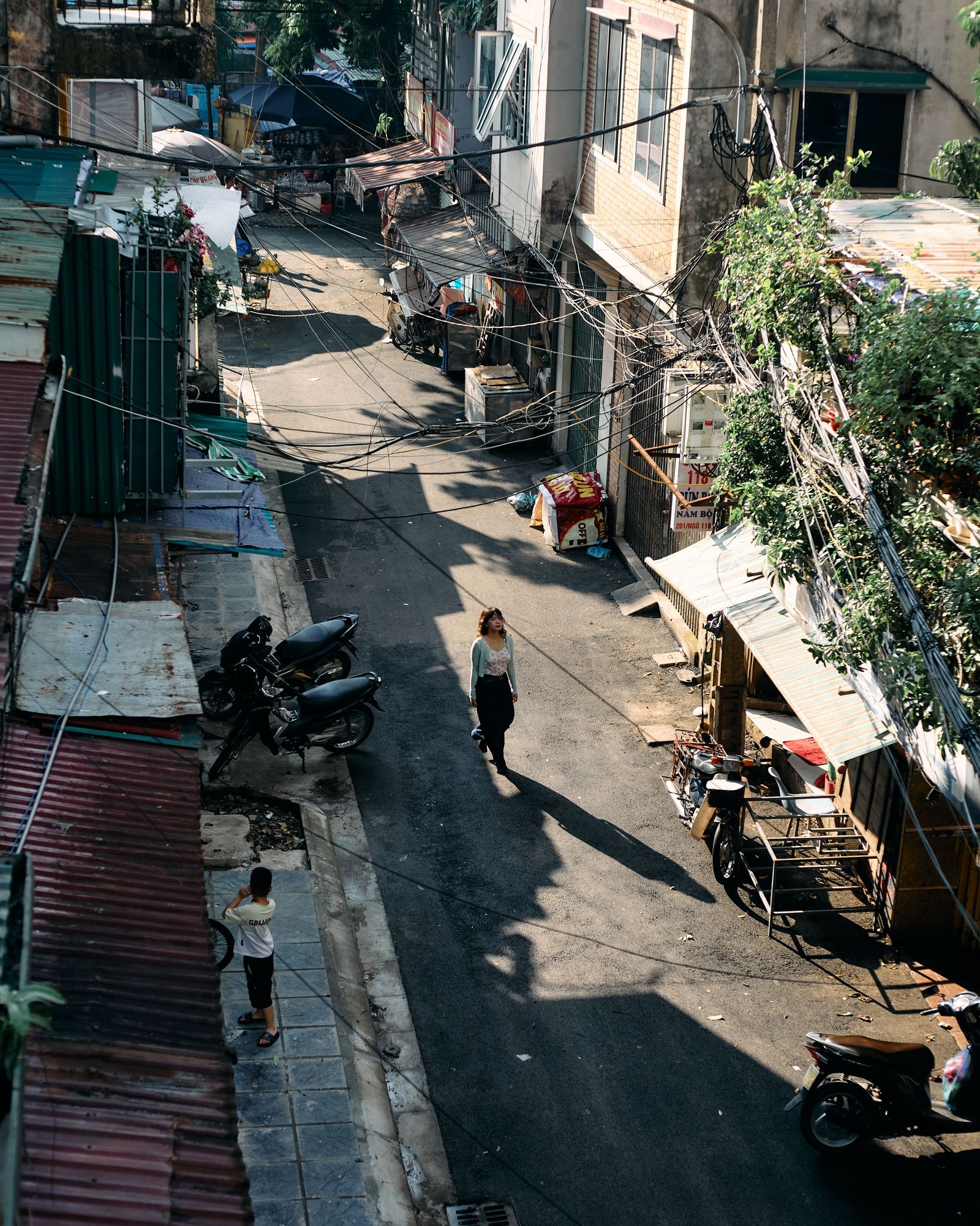 People and motorbikes move along a narrow street in Hanoi’s Old Quarter, seen from above.