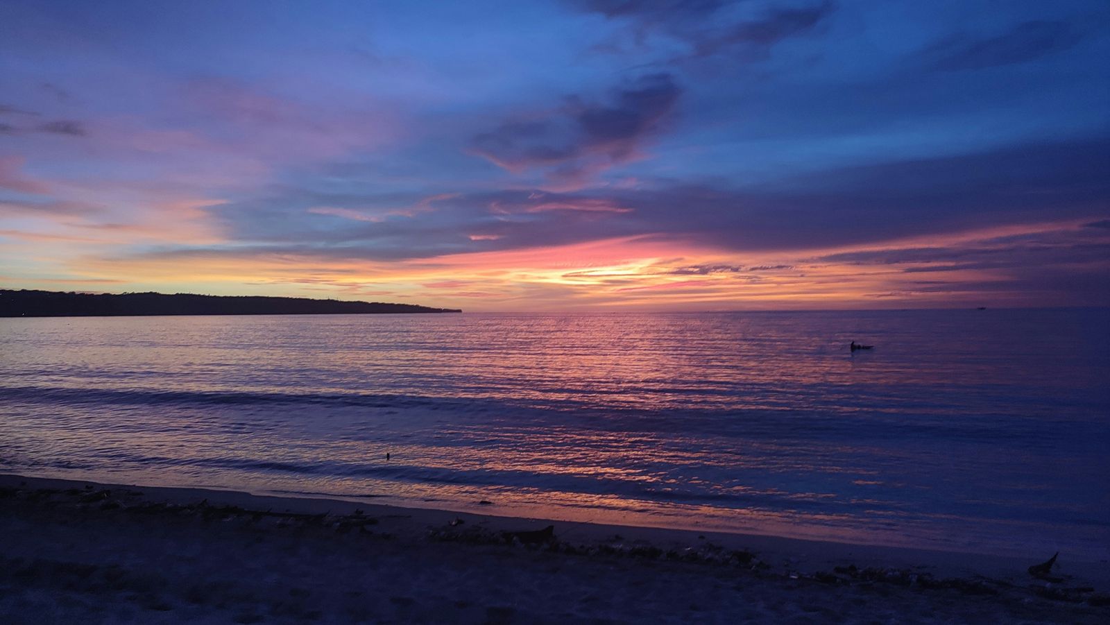 A small fishing boat sits off Jimbaran Beach beneath a pink and orange sunset.