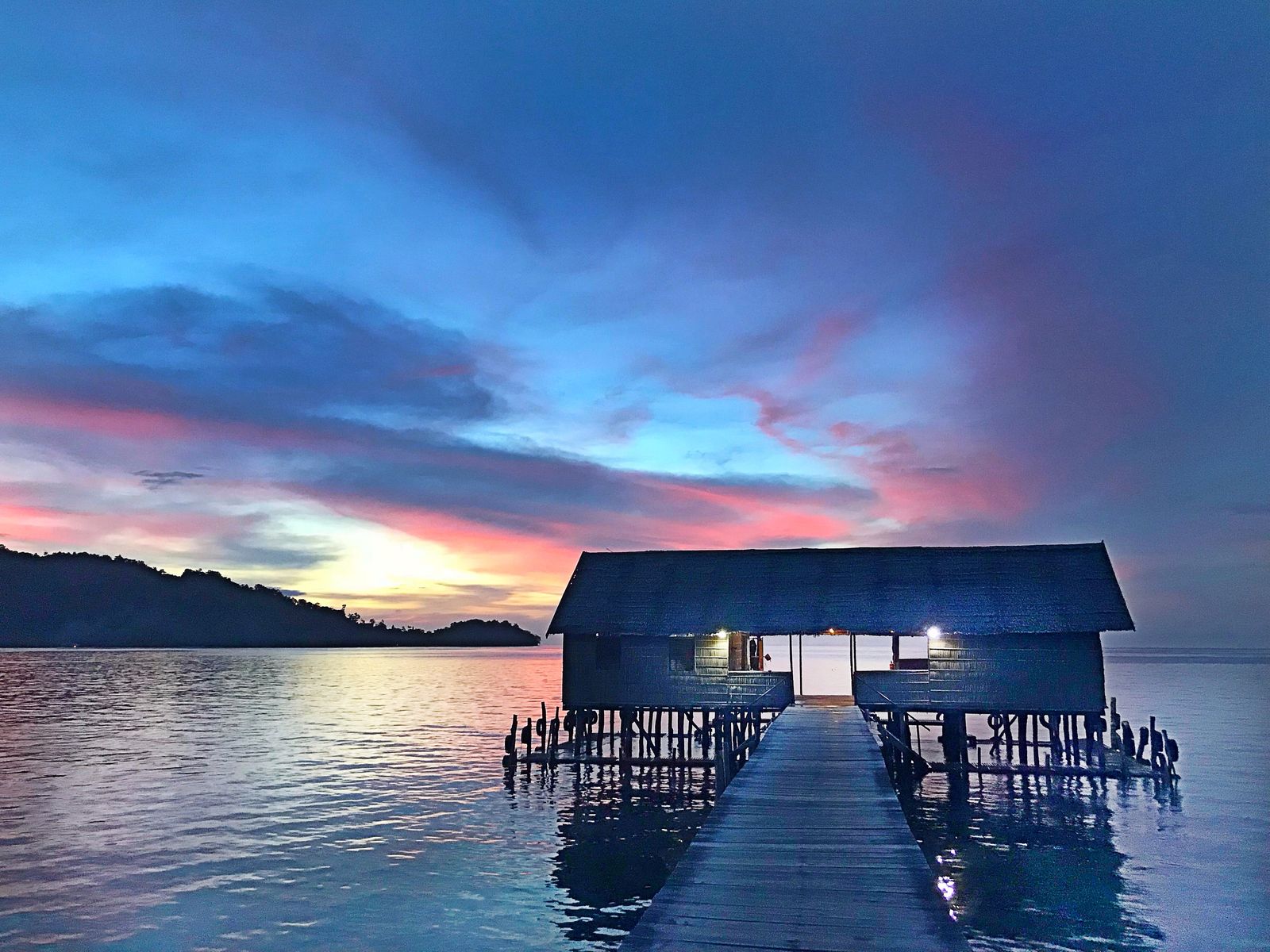 A timber jetty leads to an overwater building at Papua Paradise Eco Resort in Raja Ampat at sunset.